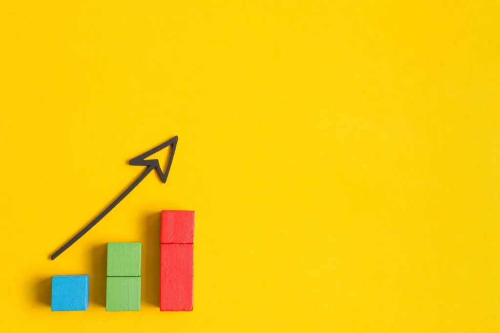 A 3D illustration of colorful bar chart cubes with a hand-drawn upward-pointing arrow on a bright yellow background, representing business growth and data success.