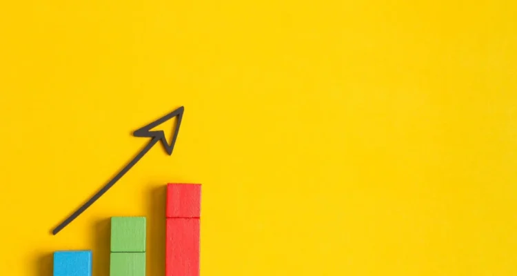 A 3D illustration of colorful bar chart cubes with a hand-drawn upward-pointing arrow on a bright yellow background, representing business growth and data success.