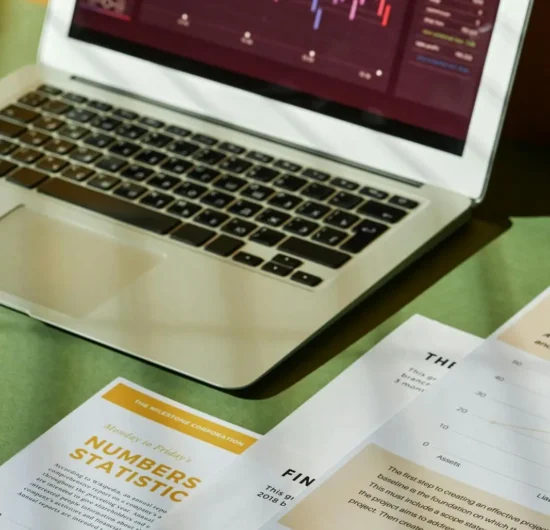 A laptop screen displaying a colorful stock market growth chart and data analytics, surrounded by business reports and financial documents on a white desk.