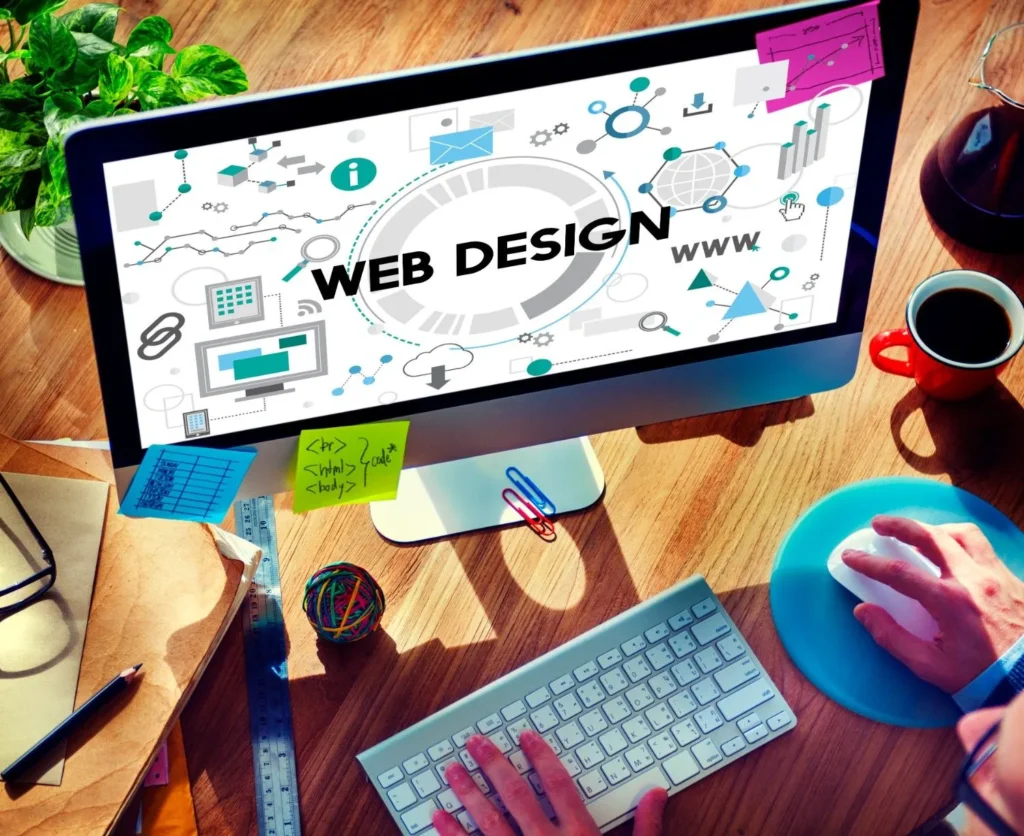A high-angle view of a workspace featuring a computer monitor displaying a "Web Design" layout, surrounded by digital design icons, a keyboard, and coffee.